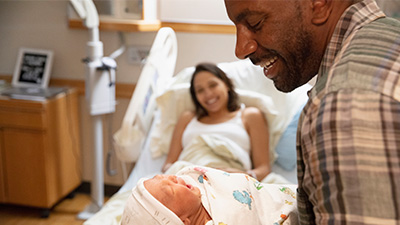 black parent holding an infant in foreground and another parent in the background