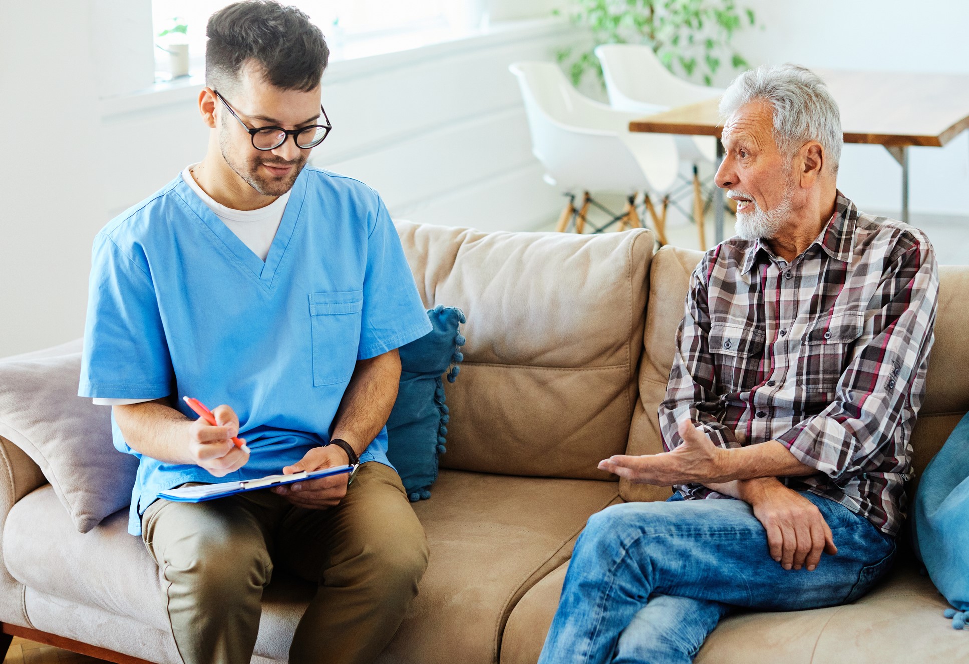 Male health worker smiles while listening to elderly client as they are seated on livingroom sofa